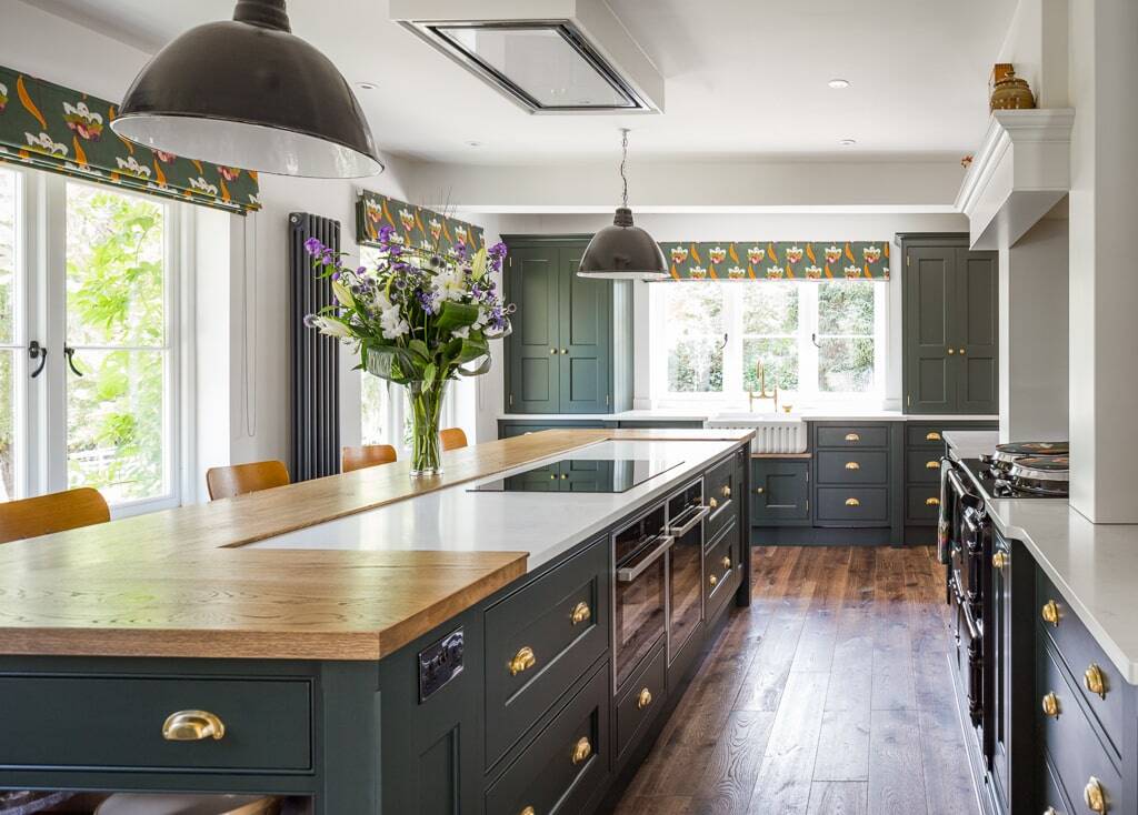 A spacious bespoke Shaker-style kitchen featuring dark green cabinetry, a large central island with a wooden worktop and integrated sink, set against a dark hardwood floor. 