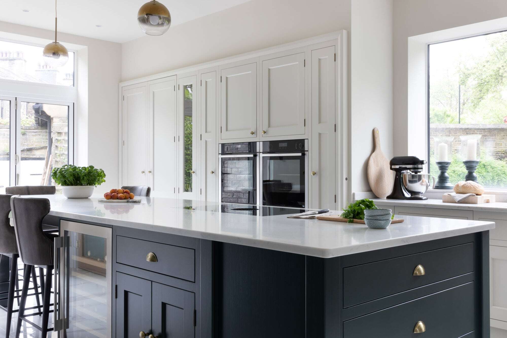 Modern and traditional kitchen design featuring a central island with dark cabinetry, sleek white countertops, a built-in double ovens and large windows allowing natural light to fill the space. 